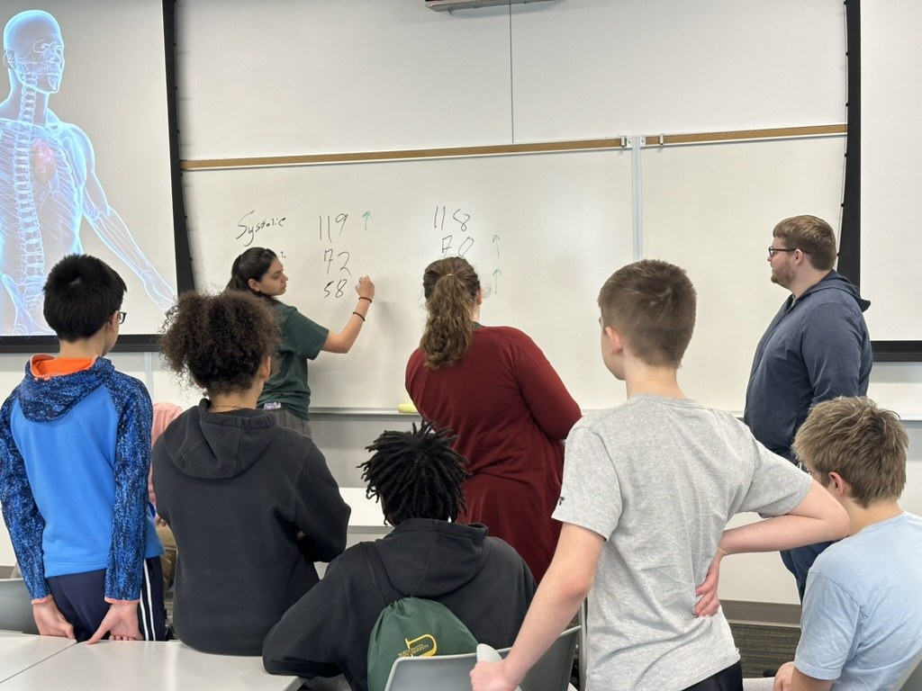 Middle and high school students watch as lab members record blood pressure and heart rate readings on a whiteboard
