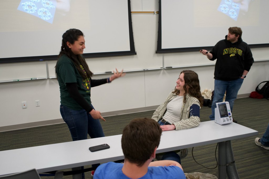 A volunteer wearing a blood pressure cuff laughs with a presenter during the Avenues 2025 demonstration.