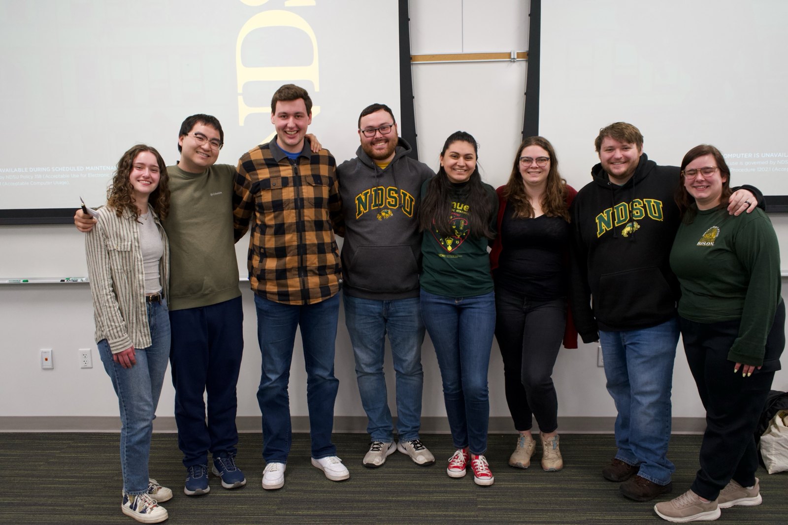 Eight members of the Avenues of Scientific Discovery 2025 team stand arm in arm in front of a projection screen.
