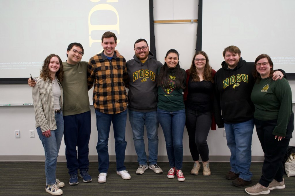 Eight members of the Avenues of Scientific Discovery 2025 team stand arm in arm in front of a projection screen.