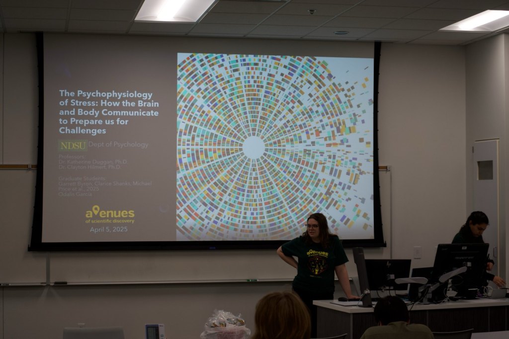 A presenter stands below the title slide: The Psychophysiology of Stress: How the Brain and Body Communicate to Prepare us for Challenges, Avenues of Scientific Discovery, April 5, 2025.
