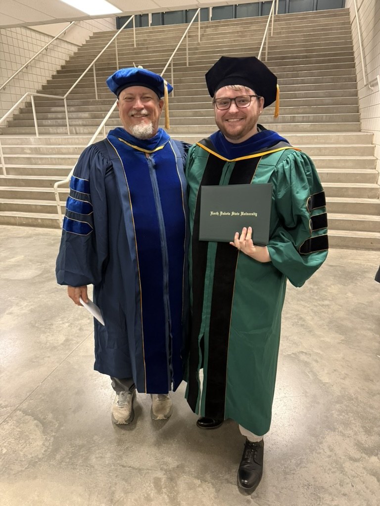 Dr. Garrett Byron holding his diploma beside Clayton Hilmert after NDSU commencement