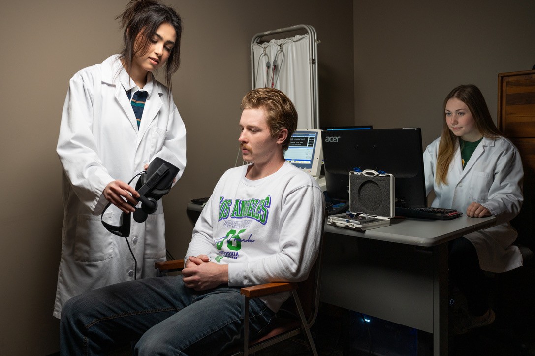A research assistant in a white lab coat shows a VR headset to a seated participant while a second research assistant in a lab coat smiles from the computer station.