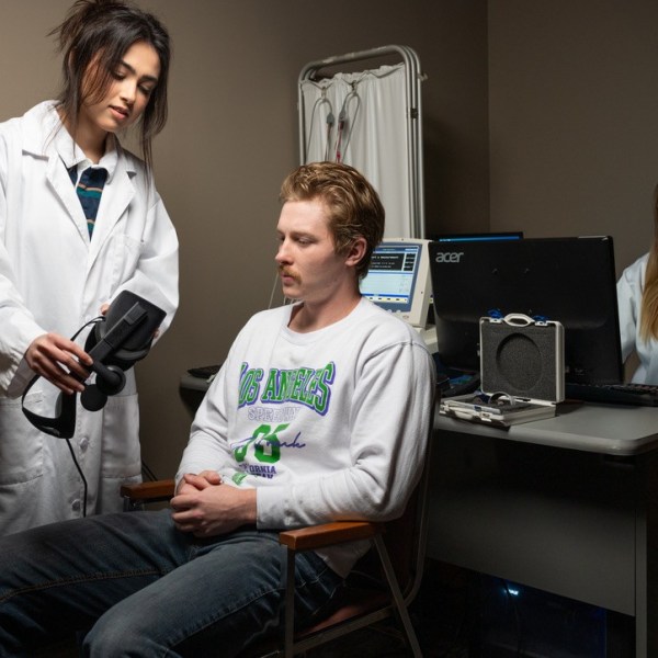 A research assistant in a white lab coat shows a VR headset to a seated participant while a second research assistant in a lab coat smiles from the computer station.