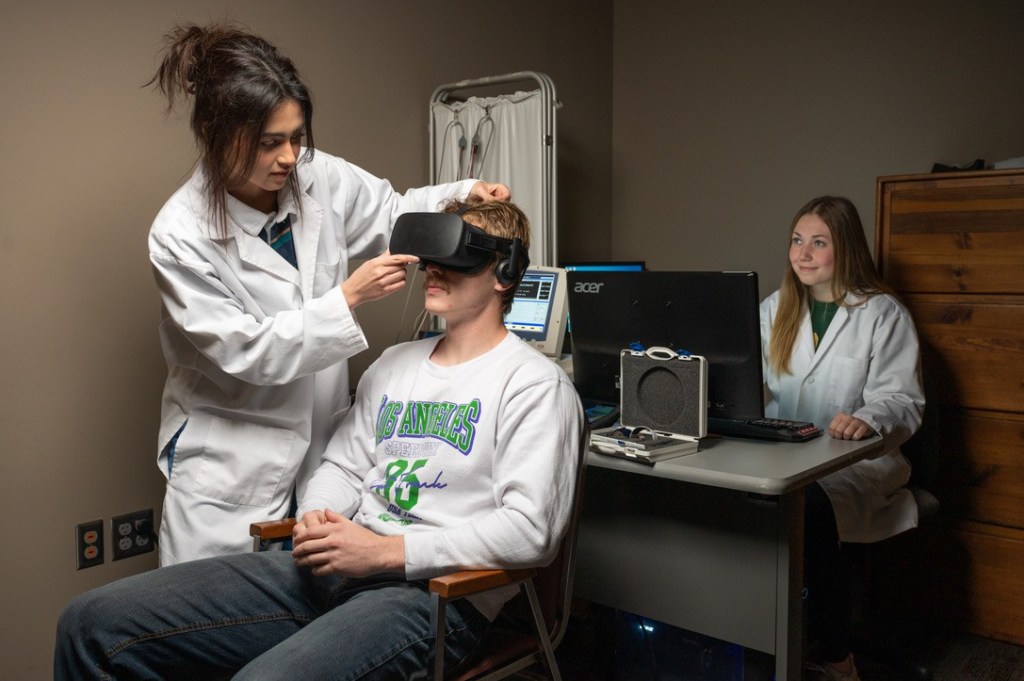 An experimenter in a white lab coat fits a VR headset on a seated participant while a second lab member in a lab coat watches from the computer station.
