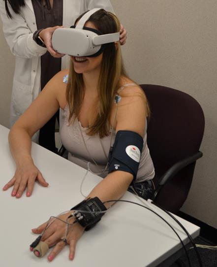 A participant wearing a VR headset, blood pressure cuff, and finger cuff sits at a lab table while an experimenter adjusts the headset.