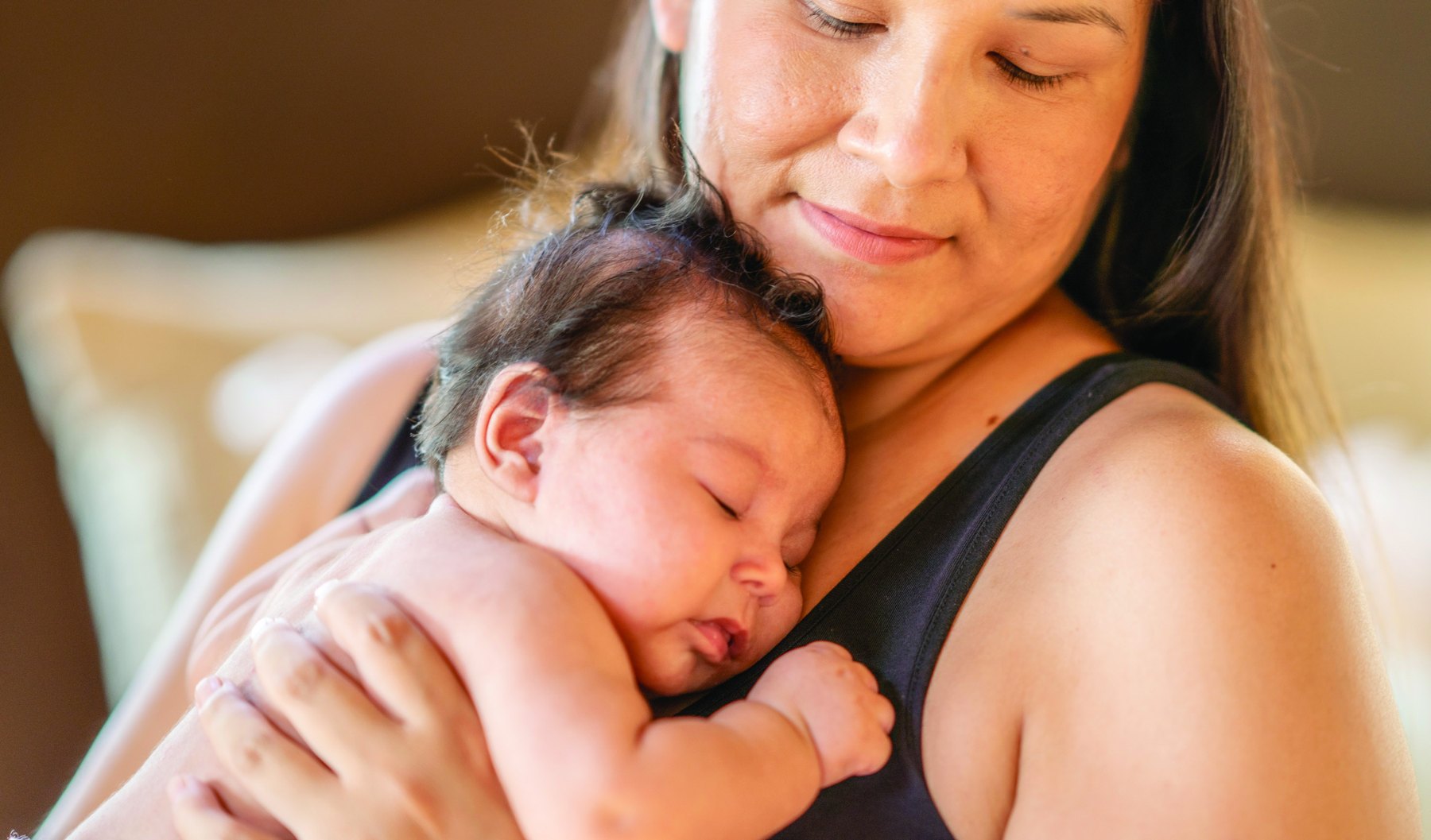 A mother smiles softly while holding her sleeping newborn against her shoulder.