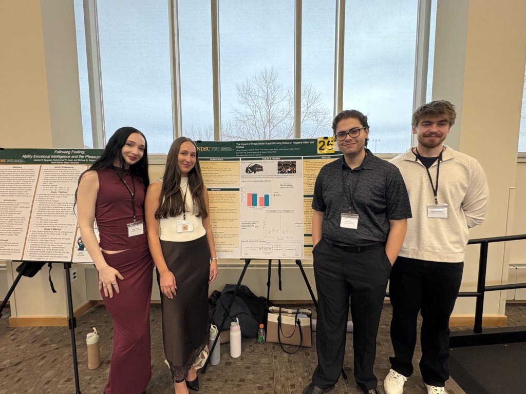 Rylie Rizzio, Ashlyn Seurer, Juan Osorno, and Trevor Cvancara stand beside their virtual social support poster at the Red River Psychology Conference 2026.