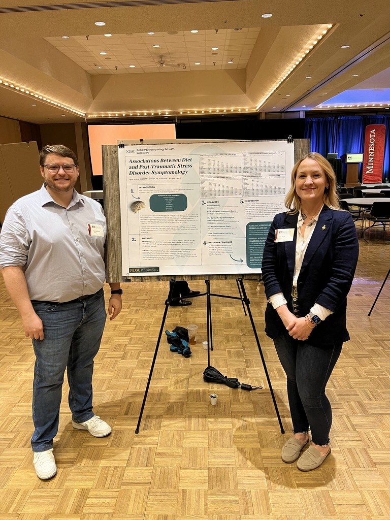 Two lab members standing beside the diet and PTSD poster at the 2024 Red River Psychology Conference