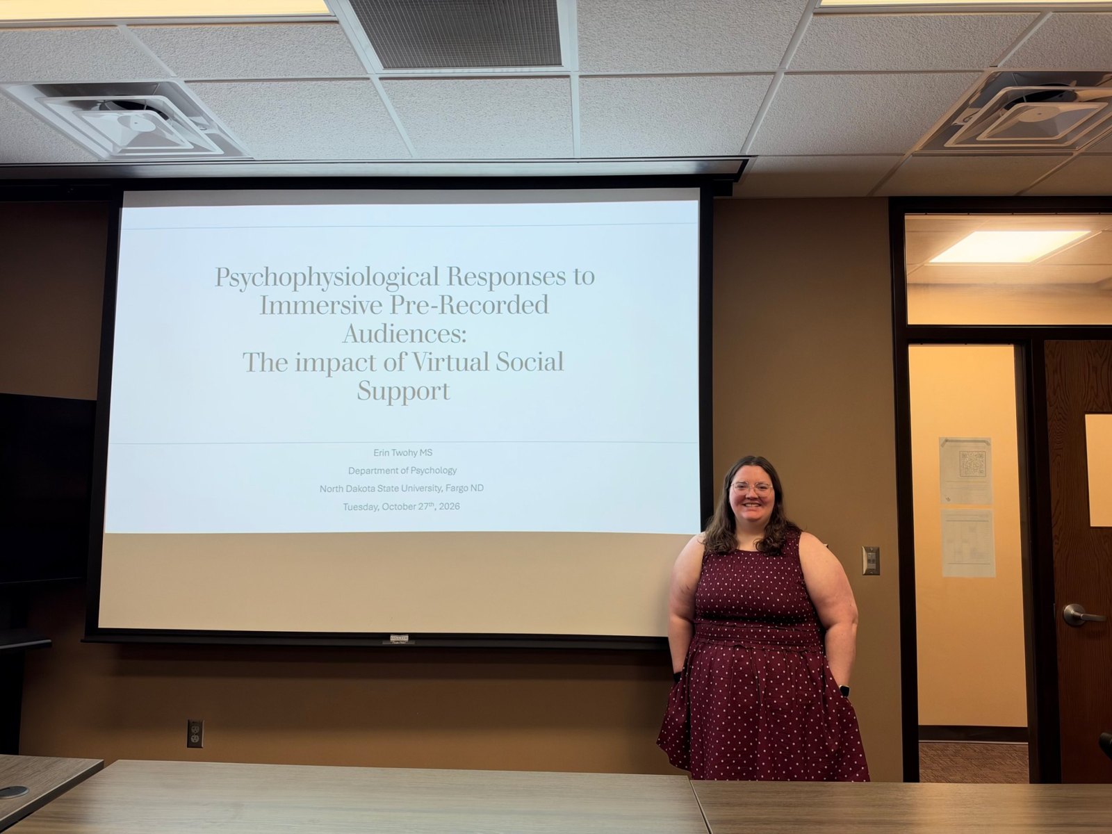 Erin Twohy smiles beside the title slide of her master's thesis defense, Psychophysiological Responses to Immersive Pre-Recorded Audiences: The Impact of Virtual Social Support.
