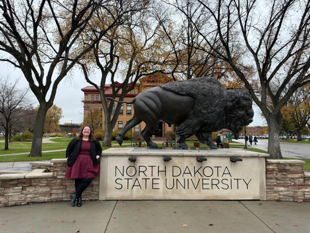 Erin Twohy smiles beside the bronze bison statue and North Dakota State University sign on campus.